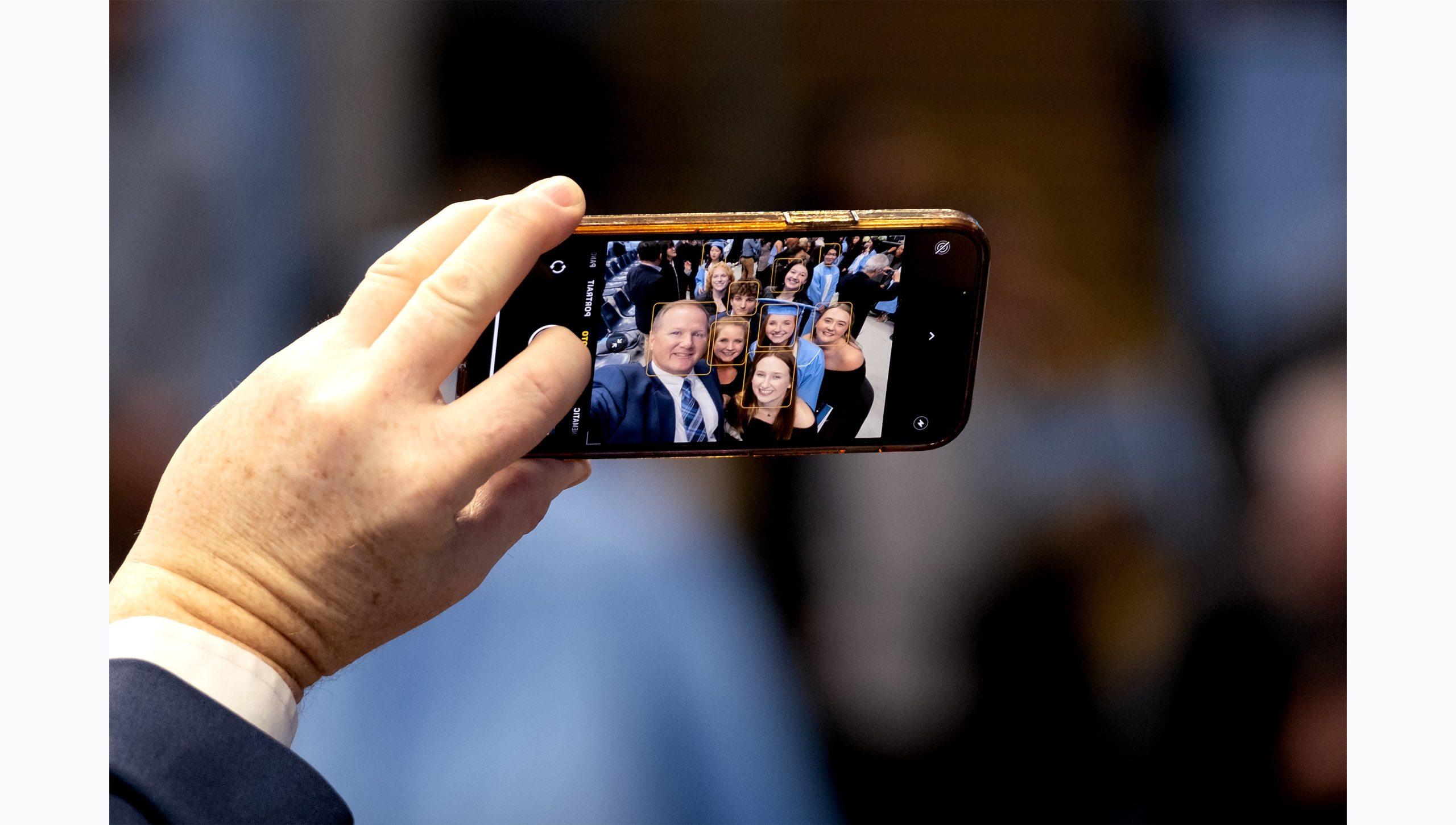 View of a hand holding up a cellphone taking a selife. Displayed on the phone screen is a family surrounding a graduate at U.N.C. Chapel Hill's Winter Commencement.